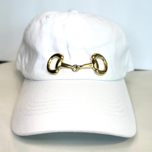 May include: A white baseball cap with a gold-colored horse bit buckle on the front.