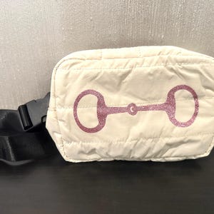 May include: A white quilted bum bag with a pink glitter horse bit design. The bag has a black adjustable strap.