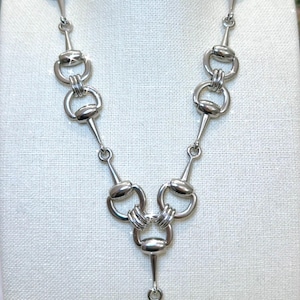 May include: Silver-toned necklace featuring a horse bit design. The necklace has a series of connected bit-shaped links, with a central pendant also in the shape of a bit. The jewelry is displayed on a white surface.