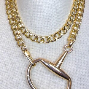 May include: Gold-toned necklace featuring two layers of chain and a large equestrian-style bit pendant. The necklace is displayed on a white surface, showcasing its shiny, reflective surface.