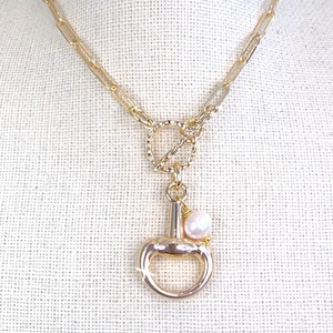 May include: Gold-toned necklace featuring a stirrup pendant with a dangling pearl. The necklace has a delicate chain and a toggle clasp. The jewelry is displayed on a neutral background.