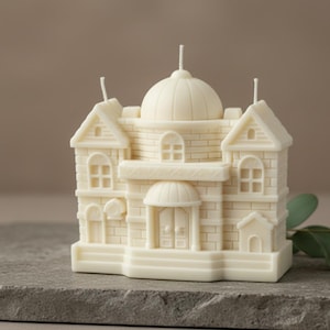 May include: Ivory-coloured candle shaped like a detailed building with a domed roof and multiple windows. The candle has a brick-like texture and three wicks. The candle sits on a grey stone surface.