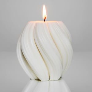 May include: A white, spiral-patterned candle with a lit wick. The candle has a glossy finish and a design of curved, vertical lines. The candle is on a reflective surface. The flame is burning brightly at the top of the candle.