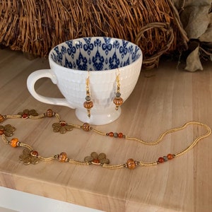 Peut inclure: Un collier en or avec des perles brunes et orange et des breloques de fleurs dorées. Le collier est présenté sur une surface en bois avec une tasse à thé blanche à motif bleu et blanc. La tasse à thé contient une paire de boucles d'oreilles dorées avec des perles brunes et grises.