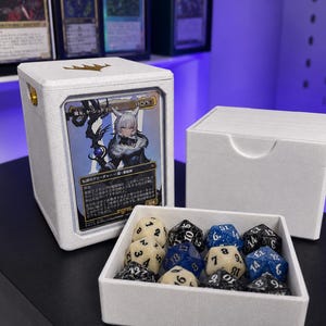 May include: A white deck box with a gold crown logo on top, displaying a trading card featuring an anime character. Next to it is a matching white tray containing nine multi-sided dice in black, cream, and blue colors with numbered faces.