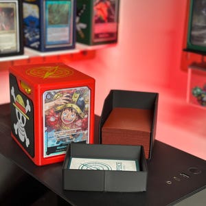 May include: A red plastic trading card deck box featuring a One Piece skull and crossbones logo on the side and a yellow lightning bolt emblem on top. The front displays a Monkey D. Luffy character card, with an open black tray holding a deck of cards.