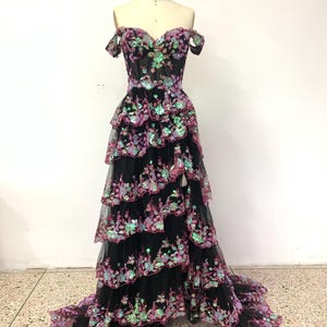 May include: A black off-the-shoulder gown with a sweetheart neckline and a tiered skirt. The skirt is adorned with iridescent pink and green sequins in a floral pattern.