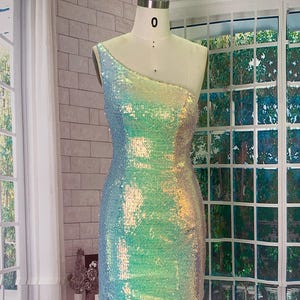 May include: A one-shoulder, iridescent sequin dress displayed on a mannequin. The dress transitions from blue to green and yellow, creating a shimmering effect. The dress is a mini-dress style.