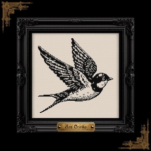 May include: A black and white cross-stitch pattern of a bird in flight, centered on a light beige fabric background. The piece is enclosed in an ornate black frame with a small gold-toned plaque at the bottom featuring the text Ars Onirika.
