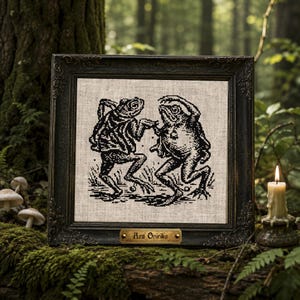 May include: A square framed cross-stitch embroidery on light beige fabric featuring a black ink-style illustration of two frogs dancing. The ornate dark frame includes a small brass-coloured plaque at the bottom with the text Ars Onirika.