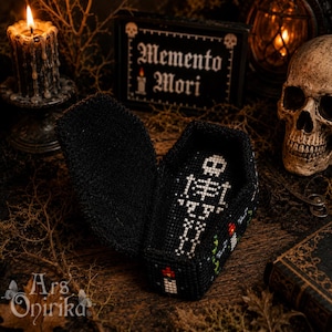 May include: A black, coffin-shaped box featuring cross-stitch embroidery. The interior base displays a white skeleton motif, while the exterior sides are decorated with small, colorful candle designs. The lid is open, revealing the detailed stitching.