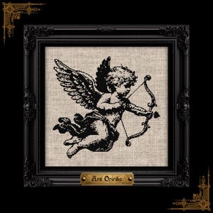 May include: A black cross-stitch embroidery of a winged cherub holding a bow and arrow, centered on a light beige linen fabric. The piece is encased in an ornate black frame with a small gold plaque at the bottom reading Ars Onirika.