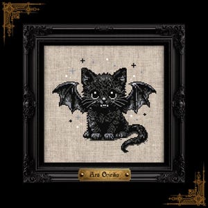 Op de afbeelding: Een kruissteekborduurwerk van een zwart kitten met vleermuisvleugels en kleine hoektanden, op een lichtbeige stoffen achtergrond. Het werk zit in een sierlijke zwarte lijst met een goudkleurig plaatje onderaan met de tekst Ars Onirika.