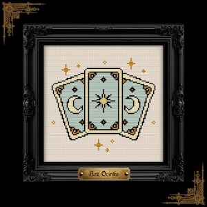 May include: A cross-stitch pattern featuring three light blue tarot cards with gold and black details, including crescent moons and a central star, surrounded by small gold stars, all displayed in a black ornate frame with a plaque reading Ars Onirika.