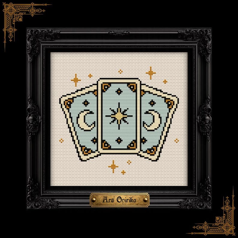 May include: A cross-stitch pattern featuring three light blue tarot cards with gold and black details, including crescent moons and a central star, surrounded by small gold stars, all displayed in a black ornate frame with a plaque reading Ars Onirika.