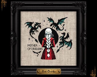 May include: A cross-stitch embroidery in a black frame featuring a person with a long white braid in a red coat, surrounded by three black and teal dragons. The text MOTHER OF DRAGONS is stitched in black, with a gold plaque reading Ars Onirika.