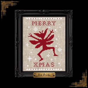 May include: A cross-stitch embroidery on light beige fabric featuring a dark red dancing creature in the center, surrounded by white stitched snowflakes. The text MERRY XMAS is stitched in dark red. The piece is enclosed in a black ornate frame.