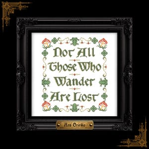 May include: Cross-stitch artwork in a black ornate frame. The piece displays the quote "Not All Those Who Wander Are Lost" in green cross-stitch. Decorative elements include mushrooms and floral accents. A small brass nameplate at the bottom reads "Ars Opirika".