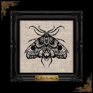 Puede incluir: Un diseño de polilla en punto de cruz negro sobre un fondo de tela beige claro, enmarcado en un marco cuadrado negro ornamentado. Una pequeña placa dorada en la parte inferior del marco presenta el texto Ars Onirika en una fuente serif oscura y estilizada.