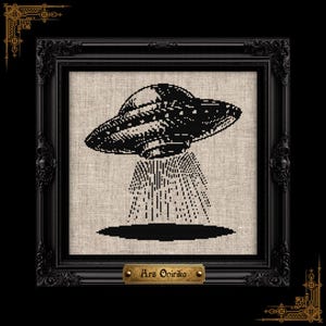 May include: A black cross-stitch embroidery of a flying saucer emitting a beam of light onto a dark oval, set against a light beige fabric background. The piece is housed in an ornate black frame with a gold plaque reading Ars Onirika.