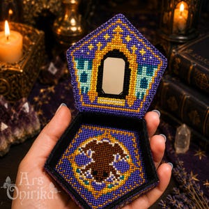 May include: A pentagonal hinged box featuring purple, gold, and teal cross-stitch embroidery. The lid displays a central mirror framed by gold stitching, while the interior base showcases a brown bat motif surrounded by a gold geometric border.