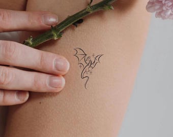 May include: A black dragon temporary tattoo design on skin. The dragon has outstretched wings, a crown, and star and crescent moon accents. The tattoo is a fine line drawing.