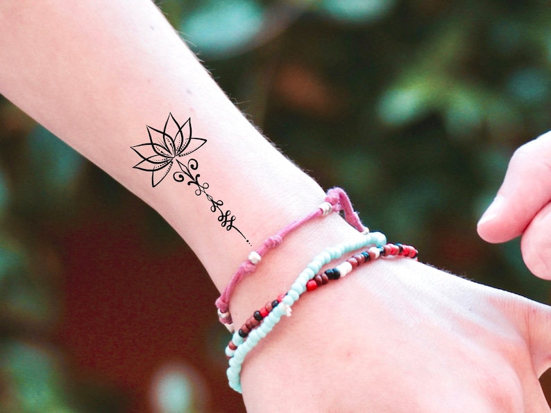 K&ouml;nnte beinhalten: Ein schwarzes und wei&szlig;es tempor&auml;res Tattoo einer Lotusblume mit einem zarten Stiel und Bl&auml;ttern. Das Tattoo befindet sich auf dem Handgelenk einer Person, die mit bunten Perlenarmb&auml;ndern geschm&uuml;ckt ist.
