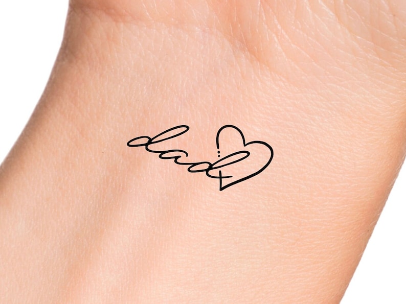 Puede incluir: Dise&ntilde;o de tatuaje negro sobre la piel. El tatuaje presenta la palabra "dad" en escritura cursiva, conectada a una forma de coraz&oacute;n. El dise&ntilde;o es simple y elegante, con una est&eacute;tica minimalista.