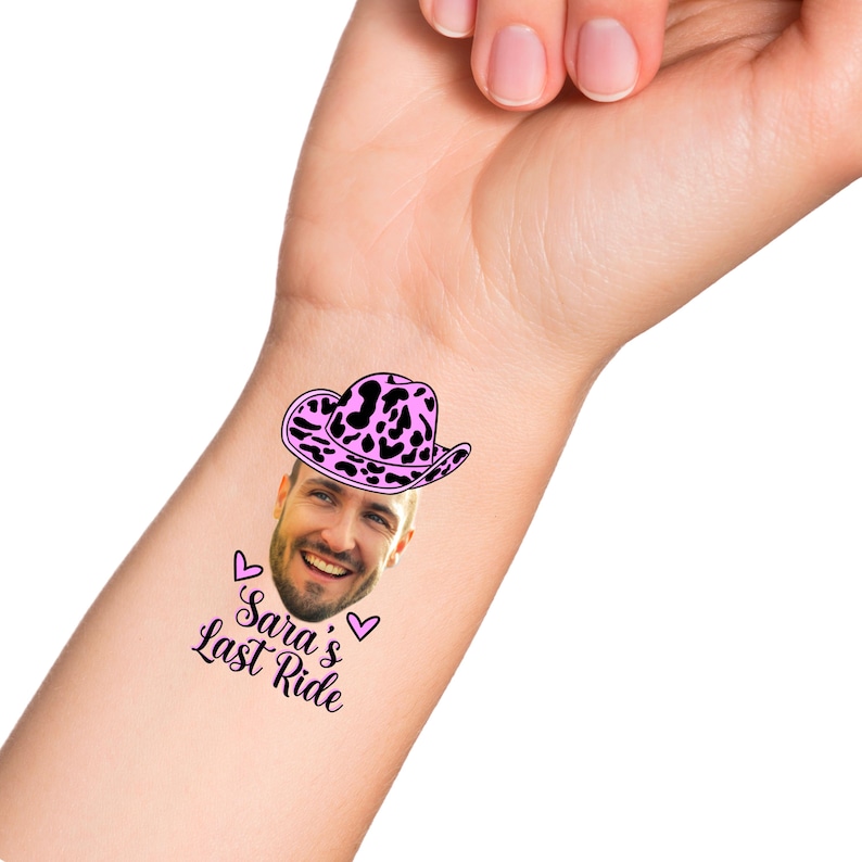 May include: A temporary tattoo with a pink and black cow print cowboy hat on a man's face. The text "Sara's Last Ride" is below the hat with two pink hearts.