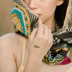 May include: A woman's hand with a small tattoo of an open book with stars coming out of it. The hand is wearing a colorful scarf.