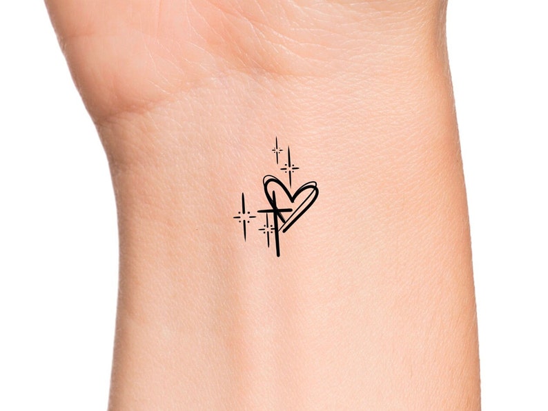 Puede incluir: Un tatuaje temporal negro con un coraz&oacute;n, una cruz y estrellas. El coraz&oacute;n est&aacute; delineado con una sola l&iacute;nea, y la cruz est&aacute; dibujada a trav&eacute;s del coraz&oacute;n. Las estrellas son peque&ntilde;as y brillantes.