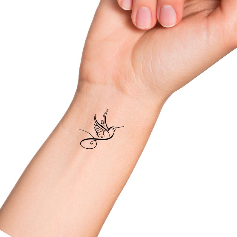 Puede incluir: Un dise&ntilde;o de tatuaje de colibr&iacute; negro en una mu&ntilde;eca. El tatuaje presenta un p&aacute;jaro estilizado en vuelo con alas detalladas y un pico largo y delgado. Una l&iacute;nea curva se extiende desde el cuerpo del p&aacute;jaro, agregando un elemento decorativo.
