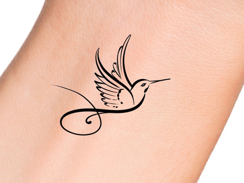 Puede incluir: Un dise&ntilde;o de tatuaje de colibr&iacute; negro. El colibr&iacute; se representa en vuelo con las alas extendidas y un pico largo y curvo. El dise&ntilde;o incluye una cola fluida y curvada. El tatuaje est&aacute; sobre un tono de piel claro.