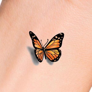 Puede incluir: Un pequeño tatuaje de mariposa naranja con marcas negras sobre un tono de piel claro.
