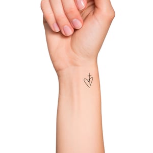 May include: A simple black line drawing of a heart with a cross inside, tattooed on a person's wrist.
