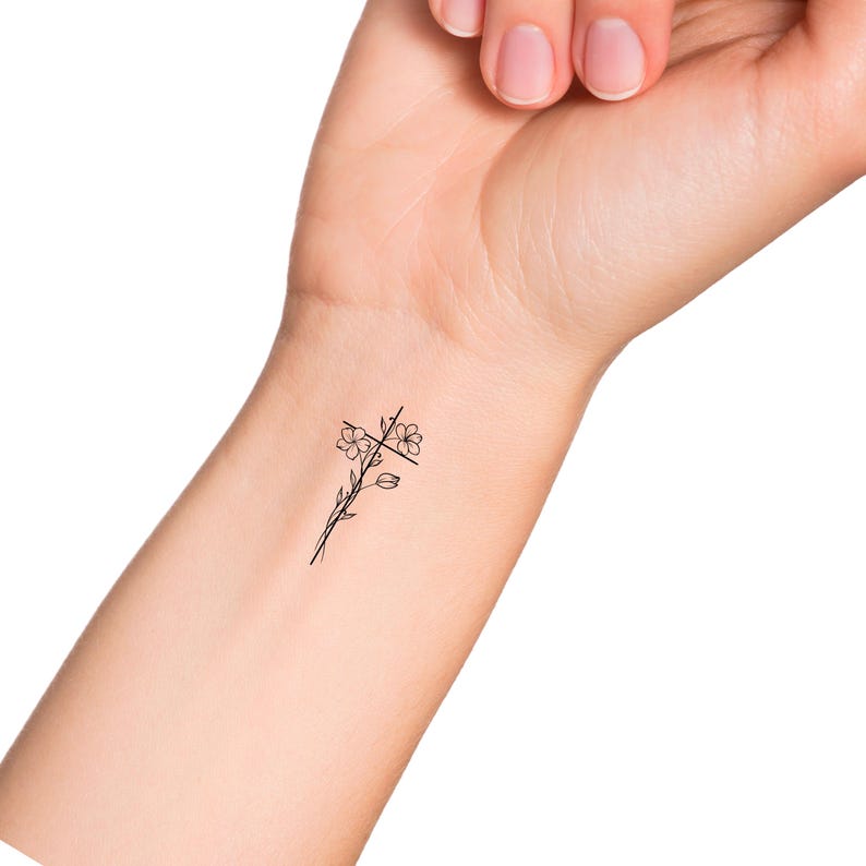 May include: A simple black line drawing of a cross with flowers on the stem. The tattoo is on a person's wrist.