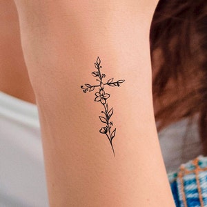 May include: A simple black line drawing of a cross with flowers and leaves. The tattoo is on a person's arm.