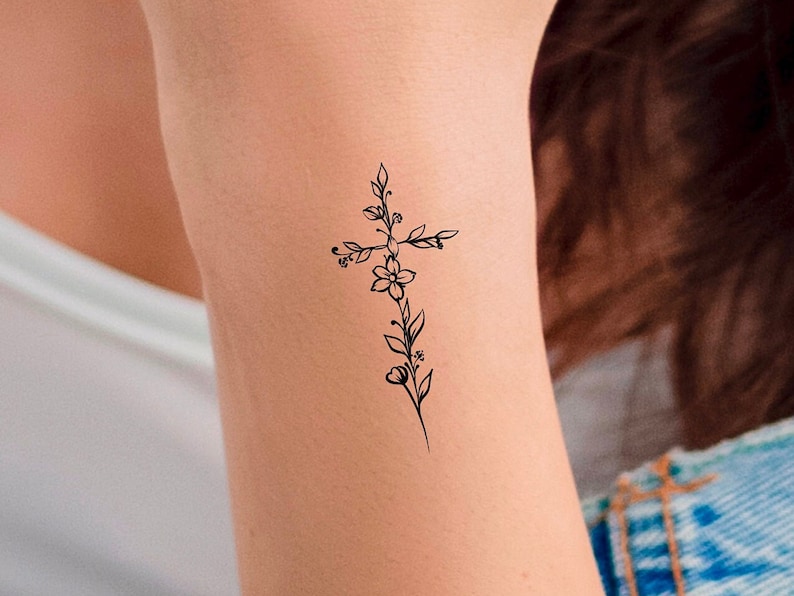 May include: A simple black line drawing of a cross with flowers and leaves. The tattoo is on a person's arm.