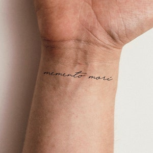 May include: A close-up of a person's wrist with a simple black tattoo that reads "memento mori".