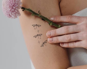 May include: Black dragonfly temporary tattoos with star accents are displayed on an arm. A pink flower and green stem are held in the hand. The tattoos are a simple, minimalist design.