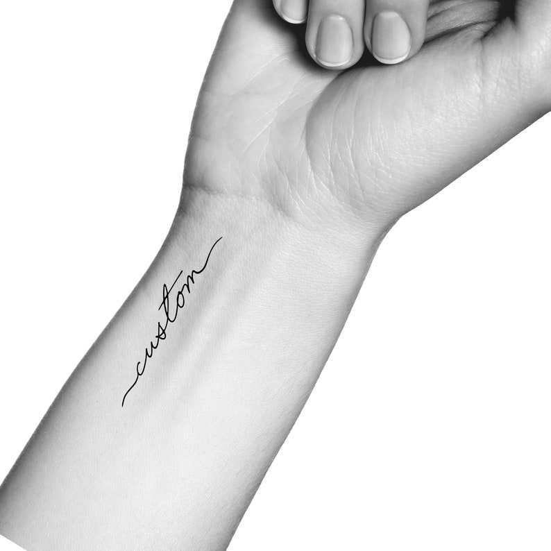 K&ouml;nnte beinhalten: Ein Schwarzwei&szlig;foto eines Handgelenks mit einem einfachen kursiv geschriebenen Tattoo, das "custom" lautet.