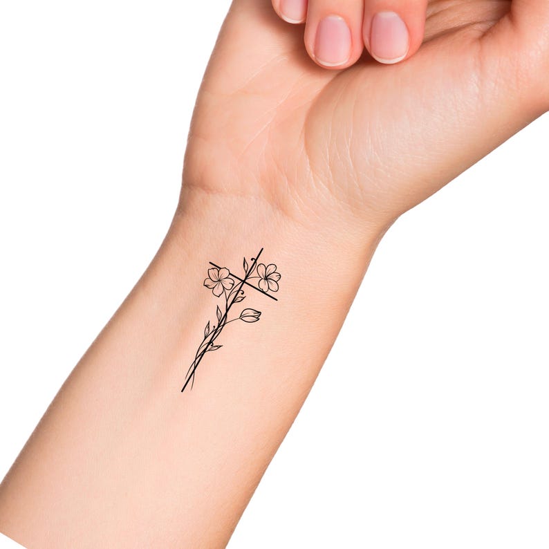 May include: A simple black line drawing tattoo design of a cross with flowers on a person's wrist.