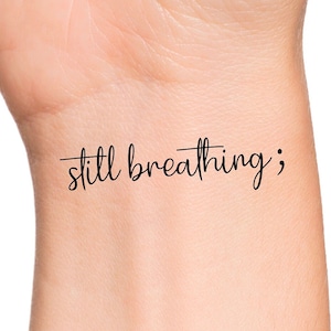 May include: A close-up of a person's wrist with a black temporary tattoo that reads "still breathing;".