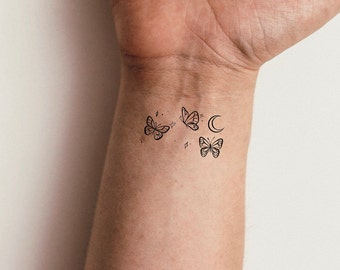 May include: A black line drawing tattoo of three butterflies and a crescent moon on a person's wrist.