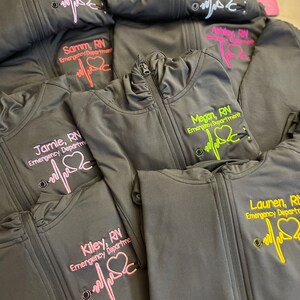May include: A stack of gray fleece jackets with embroidered names and the text "RN Emergency Department" on each jacket. The embroidery is in pink, yellow, and green thread.