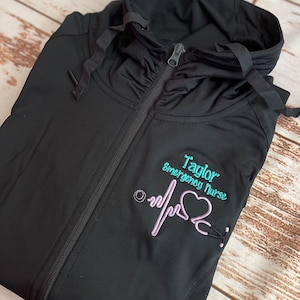 May include: Black zippered jacket with a hood. The jacket has an embroidered design on the front that says "Taylor Emergency Nurse" with a pink heart and a stethoscope.