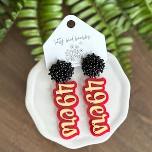 May include: Red and gold 49ers earrings with black beaded tops. The earrings are on a white plate with a small white card that says "bitty bird baubles".