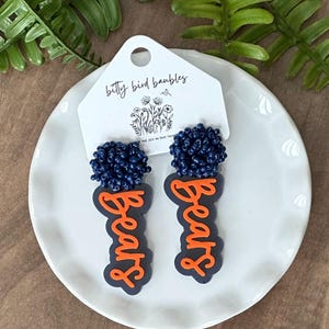 May include: A pair of navy blue beaded earrings with orange "Bears" lettering. The earrings are displayed on a white plate with a tag that reads "betty bird baubles". Green foliage is in the background.