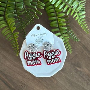 May include: A pair of earrings with the words "Aggie mom" in white script on a red background. The earrings have a silver, sparkly top and are displayed on a white, irregular-shaped plate. The background includes green foliage and a wooden surface.