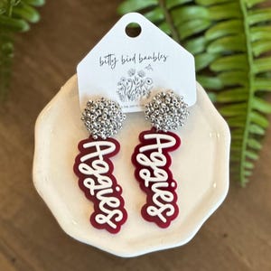 May include: Red and white acrylic earrings with the word "Aggies" written in white on a red background. The earrings are attached to silver bead clusters.
